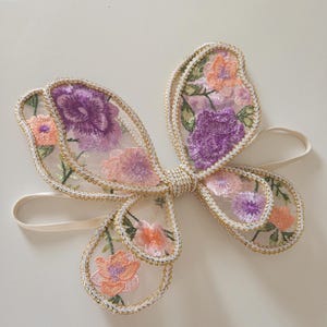 May include: A butterfly-shaped hair accessory featuring sheer mesh fabric with embroidered floral patterns in shades of purple, pink, and orange. The wings are outlined with gold metallic thread and the center is gathered with gold cord, attached to a thin elastic band.