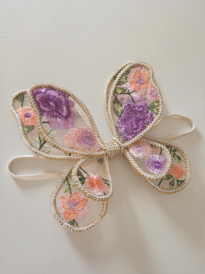 May include: A butterfly-shaped hair accessory featuring sheer mesh fabric with embroidered floral patterns in shades of purple, pink, and orange. The wings are outlined with gold metallic thread and the center is gathered with gold cord, attached to a thin elastic band.