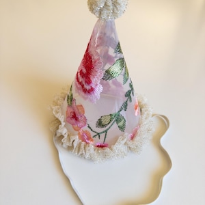 May include: A party hat with a floral design. The cone-shaped hat is made of sheer fabric and adorned with embroidered pink and red roses, green leaves, and a white beaded trim. A white pom-pom tops the hat.