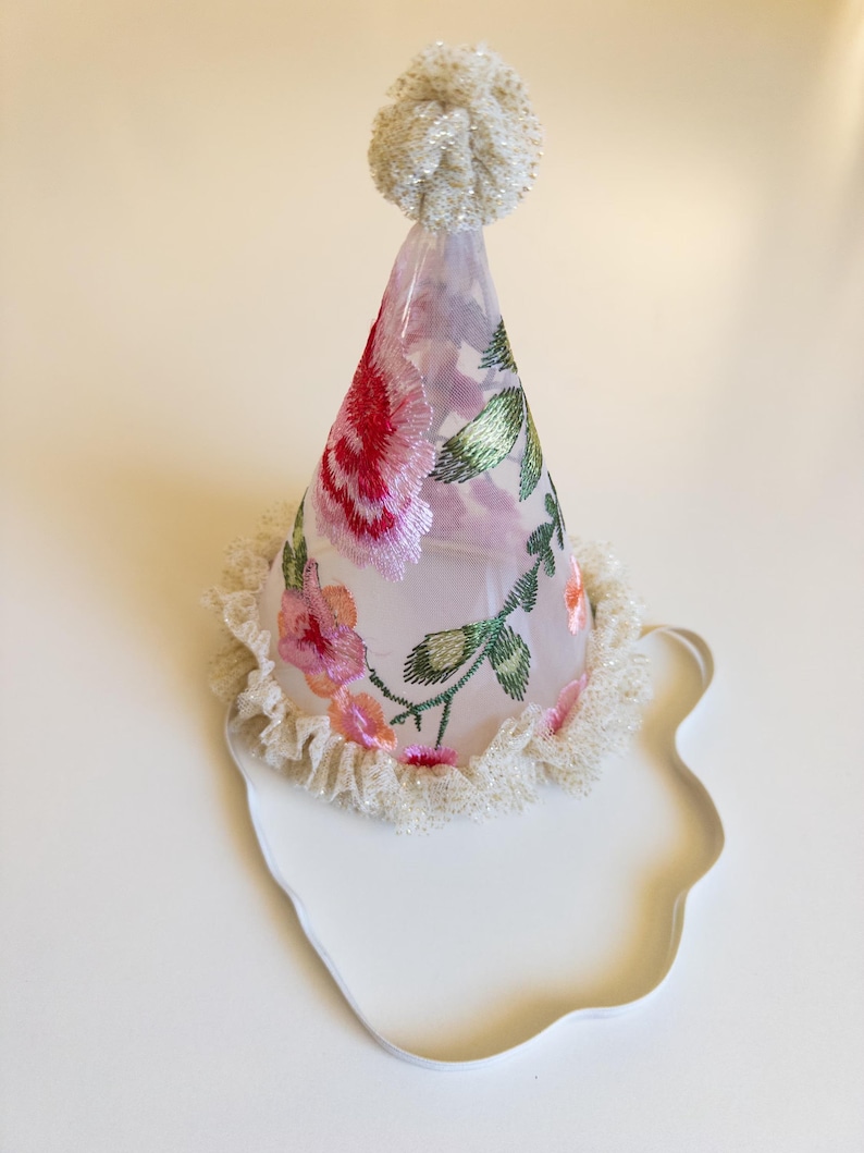 May include: A party hat with a floral design. The cone-shaped hat is made of sheer fabric and adorned with embroidered pink and red roses, green leaves, and a white beaded trim. A white pom-pom tops the hat.