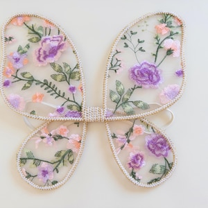 May include: Butterfly wings made of sheer, translucent fabric featuring an embroidered floral pattern in shades of purple, pink, and green. The wings are outlined with a metallic gold trim and include white elastic straps for attachment.