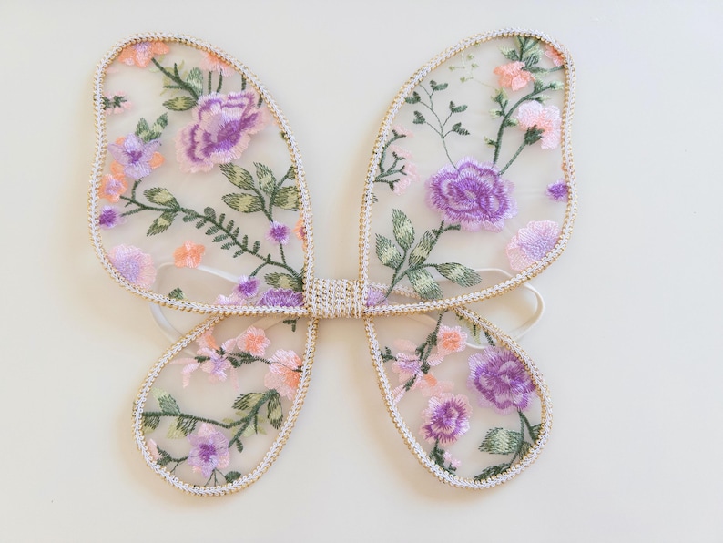 May include: Butterfly wings made of sheer, translucent fabric featuring an embroidered floral pattern in shades of purple, pink, and green. The wings are outlined with a metallic gold trim and include white elastic straps for attachment.