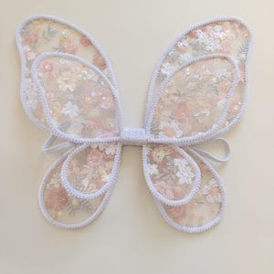 May include: A pair of butterfly wings constructed from sheer white mesh fabric featuring an intricate floral embroidery pattern in shades of soft pink, peach, and white, accented with shimmering sequins and finished with a white braided trim border.