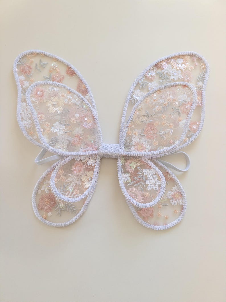 May include: A pair of butterfly wings constructed from sheer white mesh fabric featuring an intricate floral embroidery pattern in shades of soft pink, peach, and white, accented with shimmering sequins and finished with a white braided trim border.