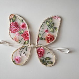 May include: Embroidered fairy wings with a floral design. The wings feature red, pink, and green floral embroidery on a sheer, cream-colored fabric. The edges are trimmed with gold beading, and a white elastic band is attached.
