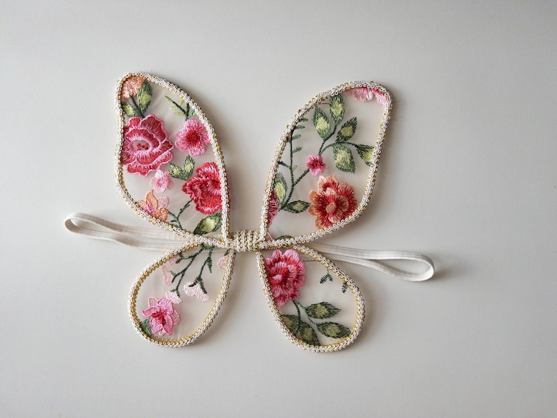 May include: Embroidered fairy wings with a floral design. The wings feature red, pink, and green floral embroidery on a sheer, cream-colored fabric. The edges are trimmed with gold beading, and a white elastic band is attached.