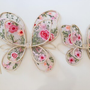 May include: A set of fairy wings and a wand. The wings are made of sheer fabric embroidered with pink and red flowers, outlined with a cream-colored trim. The wand has a pink flower on top with cream-colored ribbons.