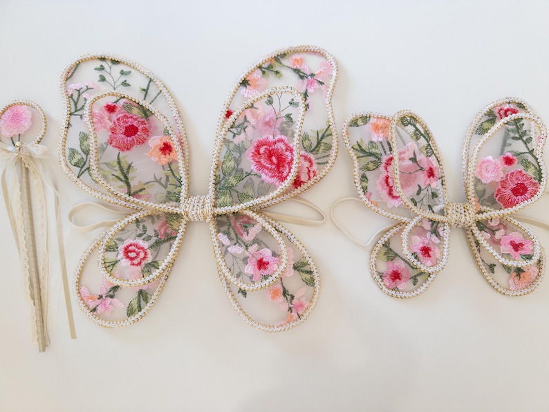 May include: A set of fairy wings and a wand. The wings are made of sheer fabric embroidered with pink and red flowers, outlined with a cream-colored trim. The wand has a pink flower on top with cream-colored ribbons.