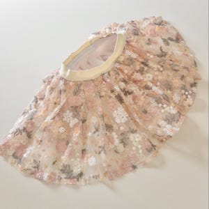 May include: A flared skirt featuring a sheer, layered tulle overlay with intricate floral embroidery in shades of soft pink, peach, and muted green. The garment includes a wide, solid cream-colored elastic waistband and a gathered, voluminous silhouette.