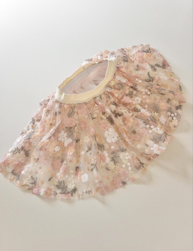 May include: A flared skirt featuring a sheer, layered tulle overlay with intricate floral embroidery in shades of soft pink, peach, and muted green. The garment includes a wide, solid cream-colored elastic waistband and a gathered, voluminous silhouette.