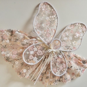 May include: A set of fairy wings and a matching skirt made from sheer, light-colored fabric featuring an intricate floral embroidery pattern in shades of pink, peach, and white. The wings have a delicate scalloped edge and a central tie closure.
