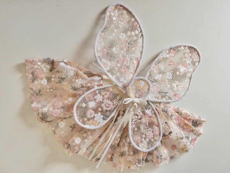 May include: A set of fairy wings and a matching skirt made from sheer, light-colored fabric featuring an intricate floral embroidery pattern in shades of pink, peach, and white. The wings have a delicate scalloped edge and a central tie closure.