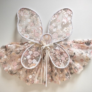 May include: A set of fairy wings and a matching skirt made from sheer fabric with an intricate floral embroidery pattern in shades of pink, white, and beige. The wings feature a white trim, and the skirt is gathered with a ribbon tie at the center.