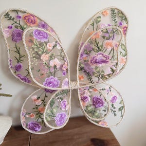 May include: A pair of butterfly wings made from sheer mesh fabric featuring intricate floral embroidery in shades of purple, peach, and green. The wings have a scalloped edge trimmed with a light-colored cord and are joined at the center.