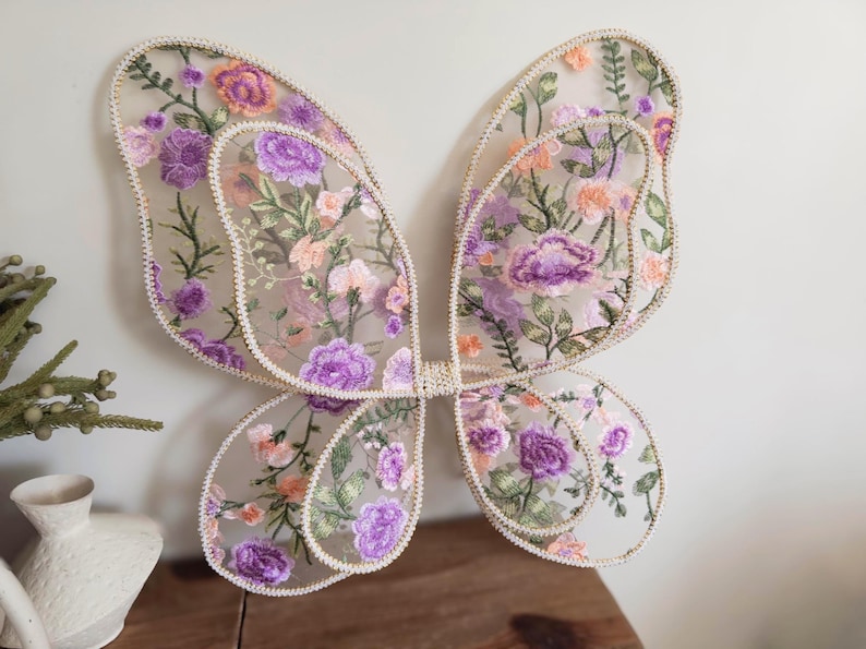 May include: A pair of butterfly wings made from sheer mesh fabric featuring intricate floral embroidery in shades of purple, peach, and green. The wings have a scalloped edge trimmed with a light-colored cord and are joined at the center.