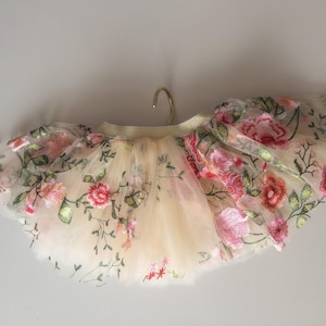 May include: A cream-colored tutu skirt adorned with embroidered pink and red flowers and green leaves. The skirt is made of layers of tulle and is hanging from a gold hanger. The floral design adds a delicate touch.