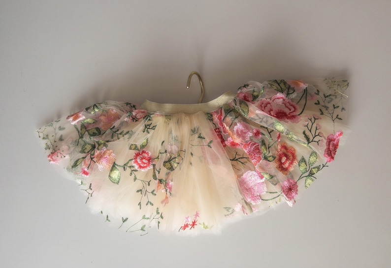 May include: A cream-colored tutu skirt adorned with embroidered pink and red flowers and green leaves. The skirt is made of layers of tulle and is hanging from a gold hanger. The floral design adds a delicate touch.