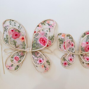 May include: Embroidered fairy wings in two sizes, with a matching wand. The wings are sheer, with a floral pattern in pink, red, and green. The edges are trimmed with a cream-colored border. The wand has a pink flower on top.