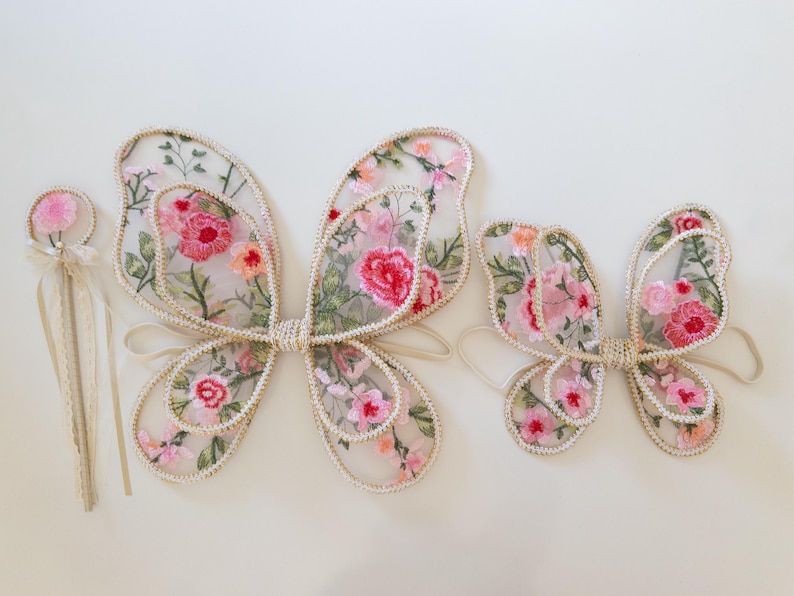 May include: Embroidered fairy wings in two sizes, with a matching wand. The wings are sheer, with a floral pattern in pink, red, and green. The edges are trimmed with a cream-colored border. The wand has a pink flower on top.