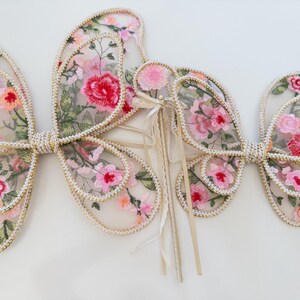 May include: Delicate butterfly wings with floral embroidery in shades of pink, red, and green. The wings are outlined with a gold trim and feature a pearl-like border. A matching wand is also included.