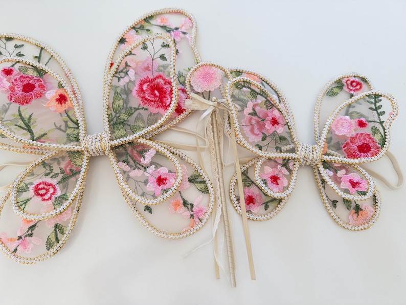 May include: Delicate butterfly wings with floral embroidery in shades of pink, red, and green. The wings are outlined with a gold trim and feature a pearl-like border. A matching wand is also included.