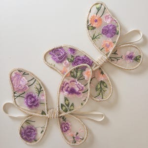 May include: Two butterfly-shaped accessories made of sheer mesh fabric with embroidered floral patterns in shades of purple, pink, and green. Each butterfly features gold-toned trim along the edges and includes a thin elastic band for attachment.