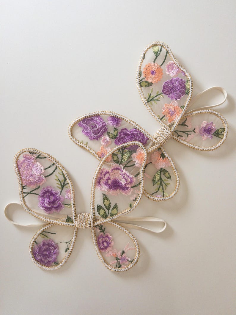 May include: Two butterfly-shaped accessories made of sheer mesh fabric with embroidered floral patterns in shades of purple, pink, and green. Each butterfly features gold-toned trim along the edges and includes a thin elastic band for attachment.