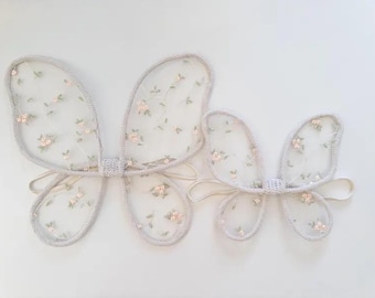 May include: Two sets of butterfly wings. The wings are made of sheer, light gray fabric with delicate floral embroidery in shades of pink and green. The edges are trimmed with a matching gray material. Each set has a matching elastic band.