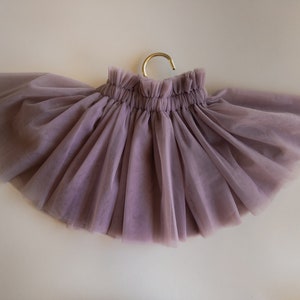 May include: A mauve tulle skirt with a gathered elastic waistband. The skirt is hanging on a gold hanger.