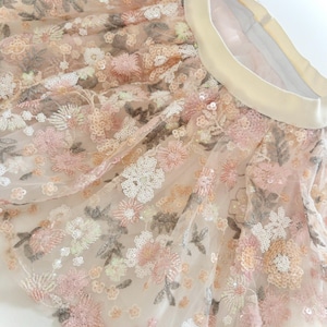May include: A tulle skirt featuring an elastic waistband and an intricate floral pattern embroidered with pink, white, and beige thread. The fabric is embellished with numerous small, shimmering sequins that catch the light across the entire surface.