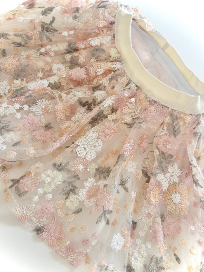 May include: A tulle skirt featuring an elastic waistband and an intricate floral pattern embroidered with pink, white, and beige thread. The fabric is embellished with numerous small, shimmering sequins that catch the light across the entire surface.
