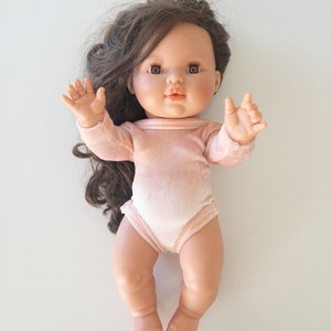 Puede incluir: Una muñeca con un body de terciopelo rosa, cabello largo castaño y ojos marrones. La muñeca lleva un body de terciopelo rosa y tiene una expresión dulce en su rostro.