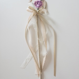 May include: A decorative wand featuring a circular top wrapped in gold cord with a small purple embroidered flower at the center. A beige organza bow with a tiny silver bell sits below the circle, with long, flowing beige lace and satin ribbons.