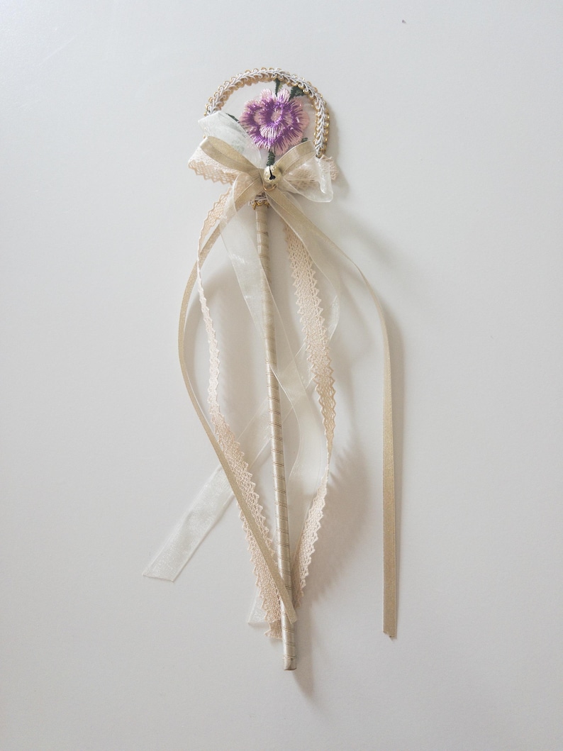 May include: A decorative wand featuring a circular top wrapped in gold cord with a small purple embroidered flower at the center. A beige organza bow with a tiny silver bell sits below the circle, with long, flowing beige lace and satin ribbons.