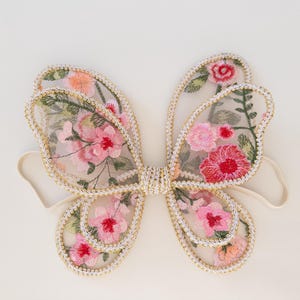 May include: Delicate fairy wings with a floral design. The wings feature a sheer, light pink fabric embroidered with pink and red flowers and green leaves. The edges are trimmed with gold-colored thread and attached to a cream-colored elastic band.