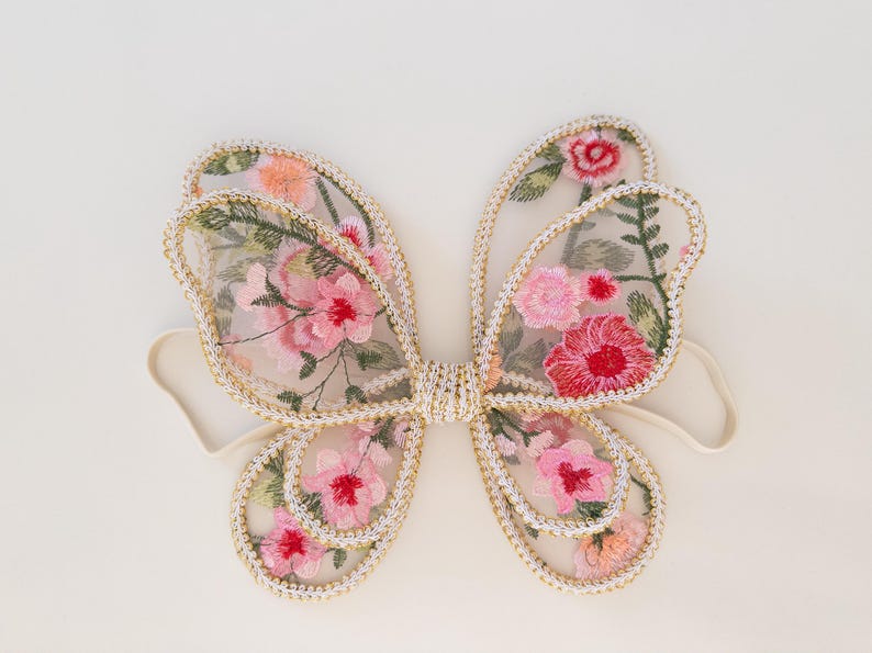 May include: Delicate fairy wings with a floral design. The wings feature a sheer, light pink fabric embroidered with pink and red flowers and green leaves. The edges are trimmed with gold-colored thread and attached to a cream-colored elastic band.