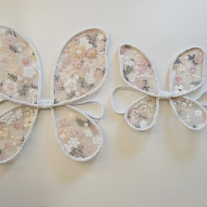 May include: Two sets of butterfly wings made from sheer white mesh fabric featuring an embroidered floral pattern in shades of pink, peach, and sage green. Each pair of wings includes white elastic arm loops for wearing as a costume accessory.