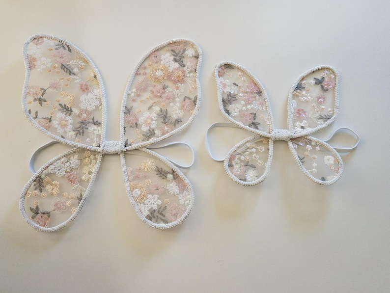 May include: Two sets of butterfly wings made from sheer white mesh fabric featuring an embroidered floral pattern in shades of pink, peach, and sage green. Each pair of wings includes white elastic arm loops for wearing as a costume accessory.
