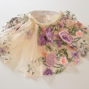 May include: A beige tulle skirt featuring an elastic waistband with a gold trim. The skirt is decorated with an intricate floral embroidery pattern in shades of purple, pink, and green, covering the sheer layered fabric throughout the garment.