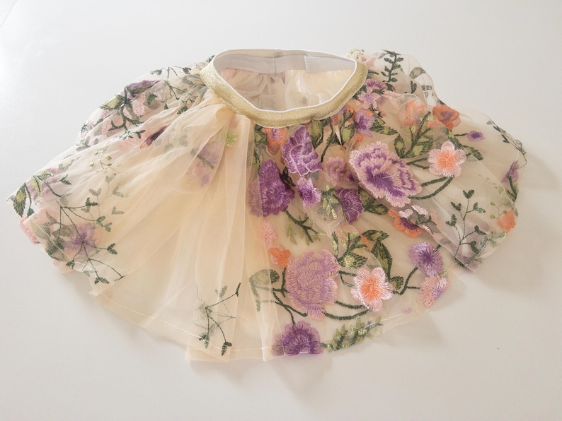 May include: A beige tulle skirt featuring an elastic waistband with a gold trim. The skirt is decorated with an intricate floral embroidery pattern in shades of purple, pink, and green, covering the sheer layered fabric throughout the garment.