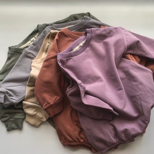 May include: Five different coloured sweatshirts stacked on top of each other. The top sweatshirt is a light purple colour, the next is a dark brown colour, the next is a light beige colour, the next is a light grey colour, and the bottom sweatshirt is a dark green colour.
