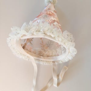 May include: A conical party hat made of sheer off-white fabric with embroidered pink floral patterns and sequins. The base features a ruffled tulle trim with silver glitter accents, a small pom-pom at the top, and an elastic chin strap.