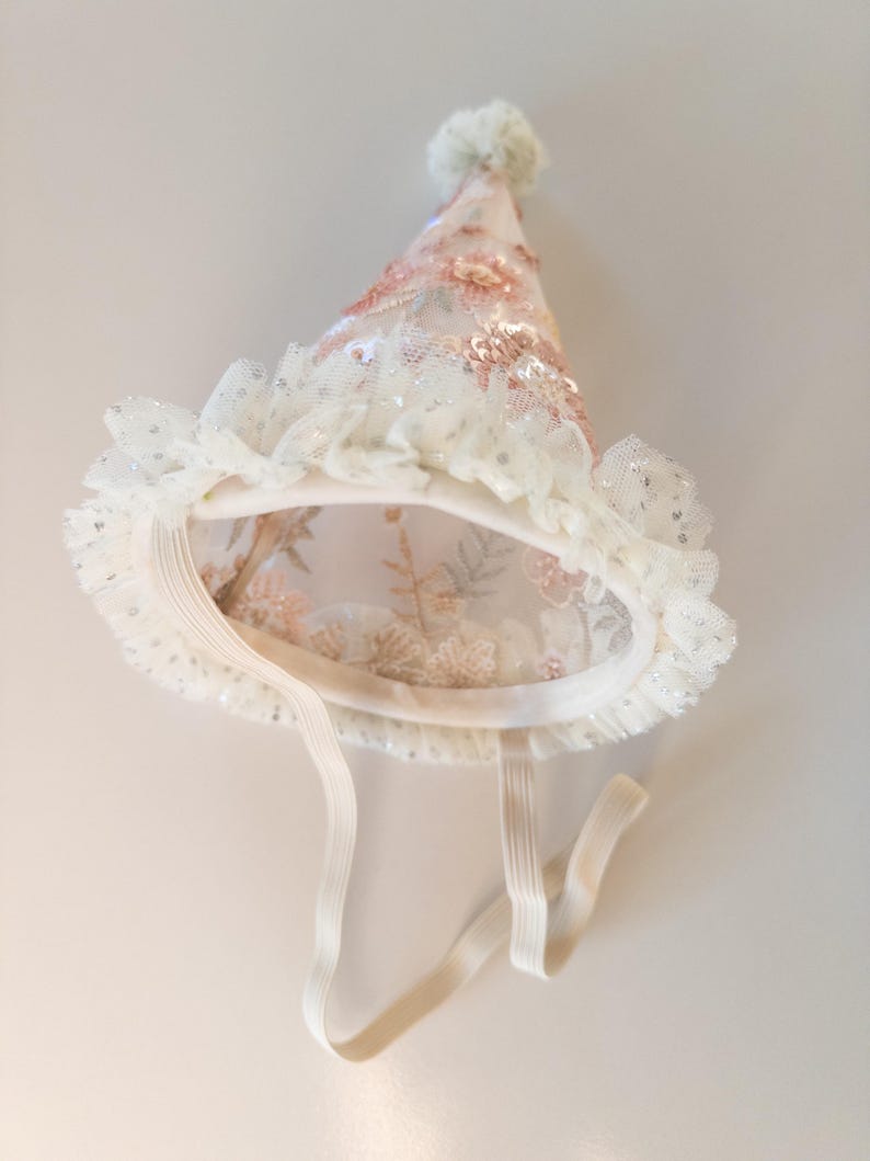 May include: A conical party hat made of sheer off-white fabric with embroidered pink floral patterns and sequins. The base features a ruffled tulle trim with silver glitter accents, a small pom-pom at the top, and an elastic chin strap.
