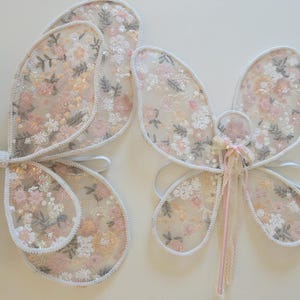 May include: A set of butterfly wings and a matching wand featuring a sheer, light-colored fabric with an intricate floral pattern in shades of pink, peach, and white. The wings have a white trim and elastic straps, while the wand has a pink handle.