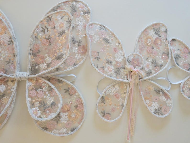 May include: A set of butterfly wings and a matching wand featuring a sheer, light-colored fabric with an intricate floral pattern in shades of pink, peach, and white. The wings have a white trim and elastic straps, while the wand has a pink handle.