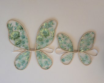 May include: Two sets of fairy wings. The wings are made of sheer, light blue fabric with green floral embroidery and gold trim. The wings are connected by a beige-colored center and have white elastic straps.