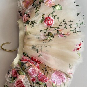 May include: Ivory tulle fabric with embroidered floral design. The embroidery features pink, red, and green flowers and leaves. The fabric is gathered and attached to a gold elastic band.