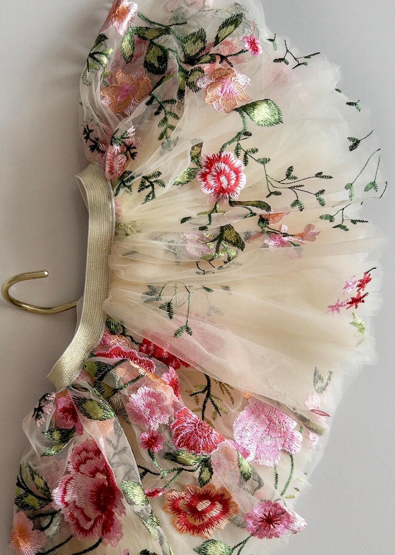 May include: Ivory tulle fabric with embroidered floral design. The embroidery features pink, red, and green flowers and leaves. The fabric is gathered and attached to a gold elastic band.
