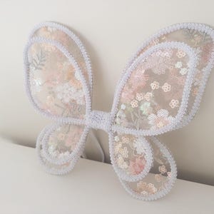 May include: A pair of butterfly wings made from sheer, light-colored fabric featuring an intricate floral pattern in soft pink and white tones. The wings are embellished with shimmering sequins and finished with a white braided trim along the edges.