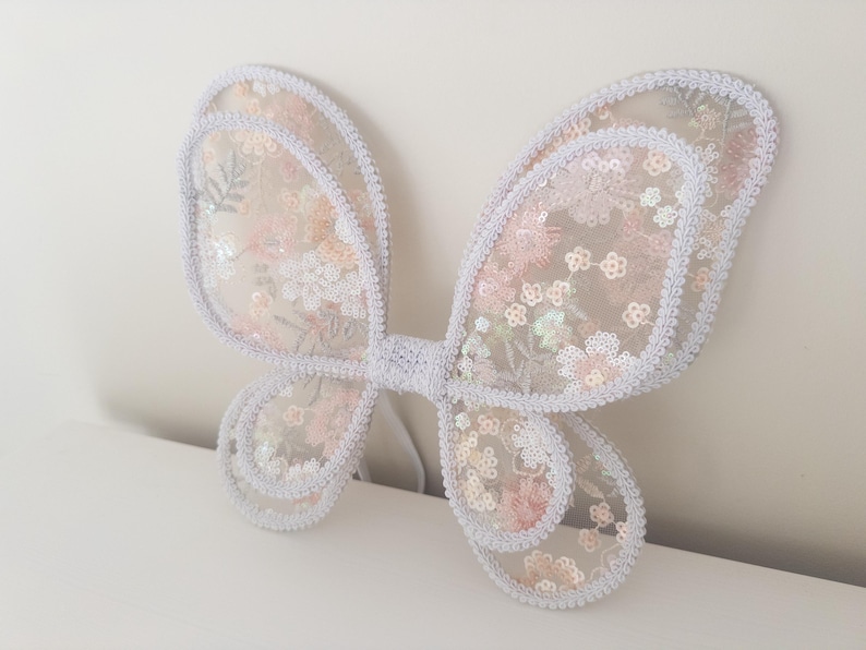 May include: A pair of butterfly wings made from sheer, light-colored fabric featuring an intricate floral pattern in soft pink and white tones. The wings are embellished with shimmering sequins and finished with a white braided trim along the edges.